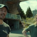 "Two people stand outdoors near modern industrial buildings on a sunny day. The man wears a cap and casual clothes, while the woman stands with her hands on her hips, both looking in the same direction."