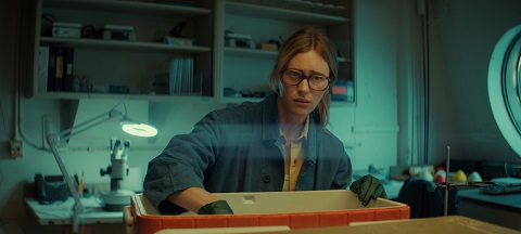"A woman wearing glasses and gloves works in a laboratory, reaching into a container on a counter, surrounded by scientific equipment and shelves in a well-lit room."